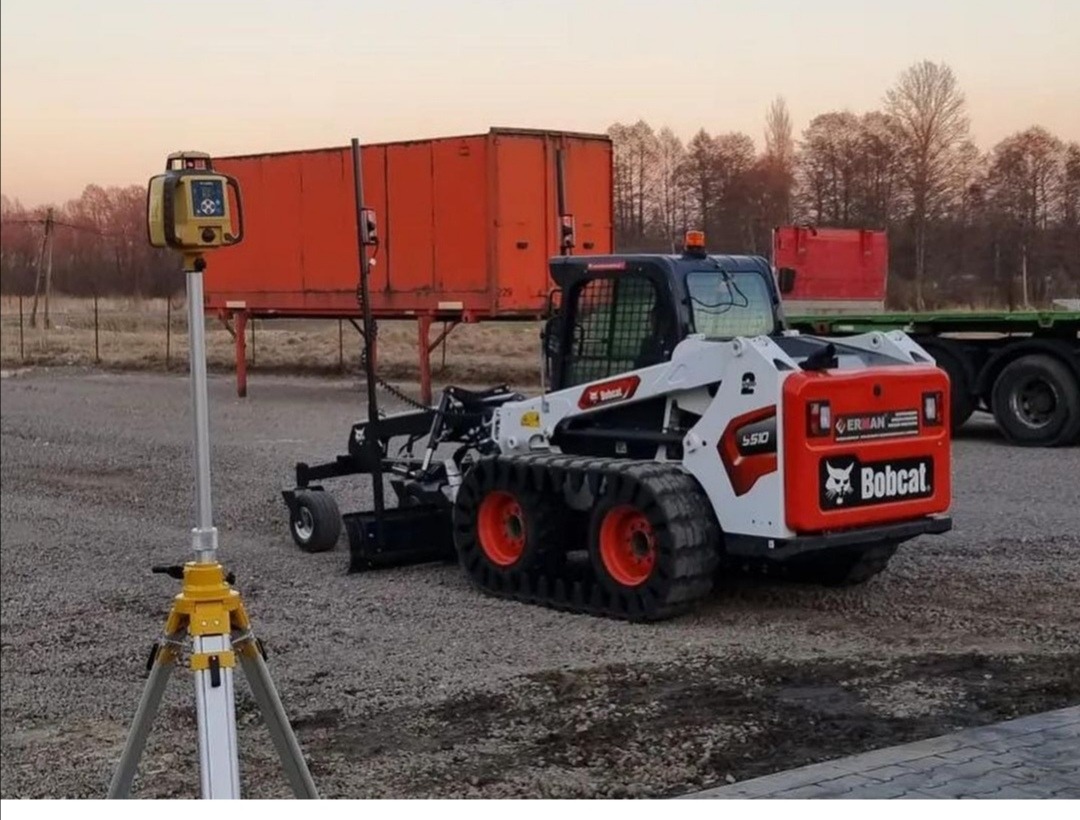 Niwelator laserowy na statywie obok białej miniładowarki gąsienicowej Bobcat S510 z osprzętem do niwelacji terenu, w tle pomarańczowa naczepa.