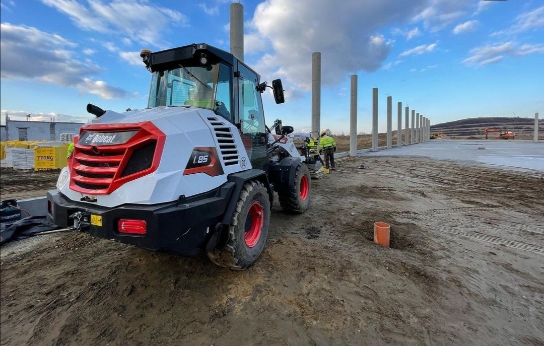 Bobcat I.85 na placu budowy z betonowymi słupami w tle, pracownik zagęszcza grunt wibratorem, widoczna sterta bloczków YTONG.