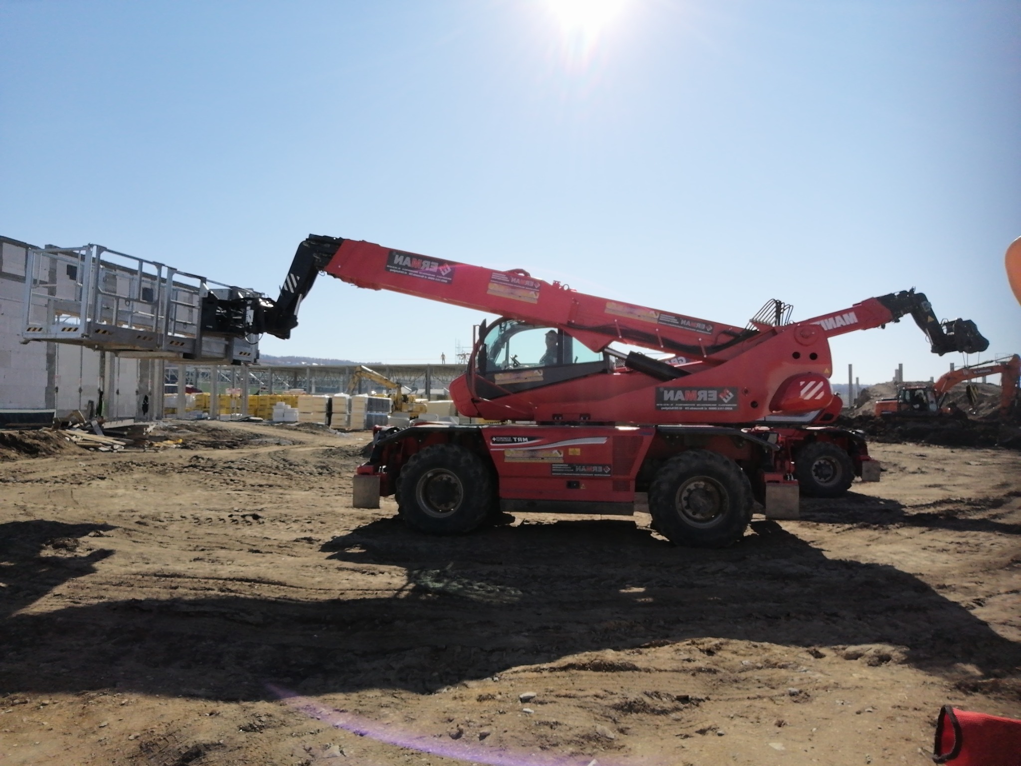 Czerwony podnośnik teleskopowy MANITOU z platformą roboczą podnosi prefabrykowany element ściany na placu budowy w słoneczny dzień. W tle widoczne inne maszyny budowlane i materiały.