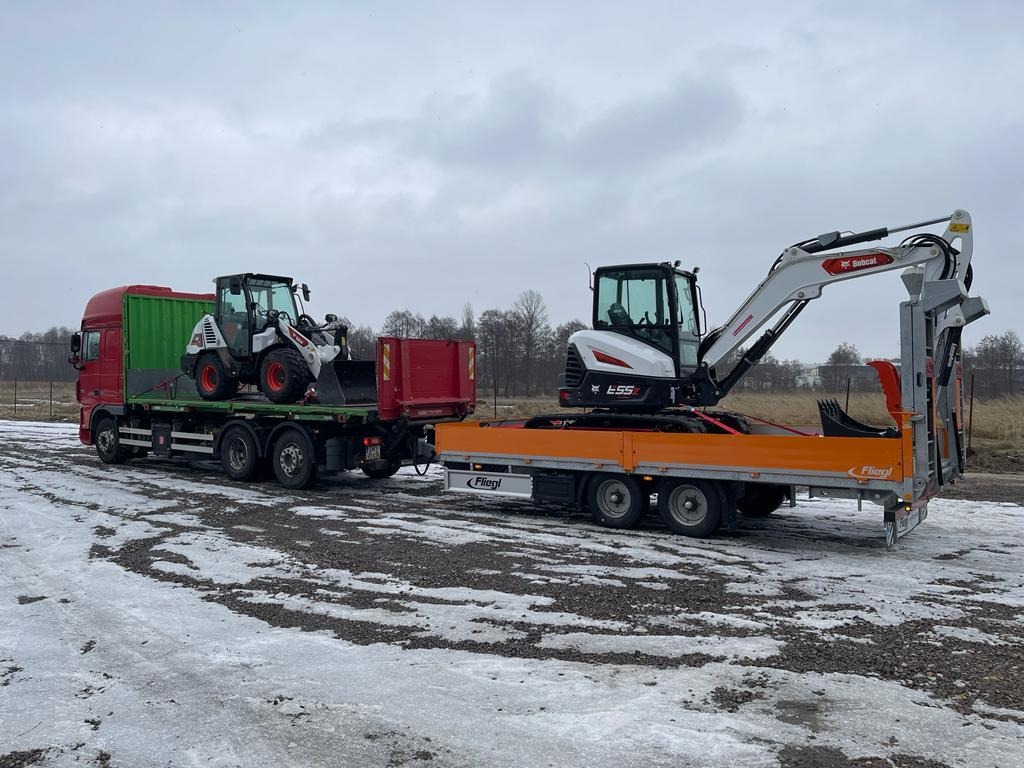 Czerwona ciężarówka z zielonym kontenerem przewozi na platformie ładowarkę Bobcat, a na pomarańczowej przyczepie koparkę Bobcat E55; zimowy krajobraz z resztkami śniegu na ziemi i pochmurnym niebem...