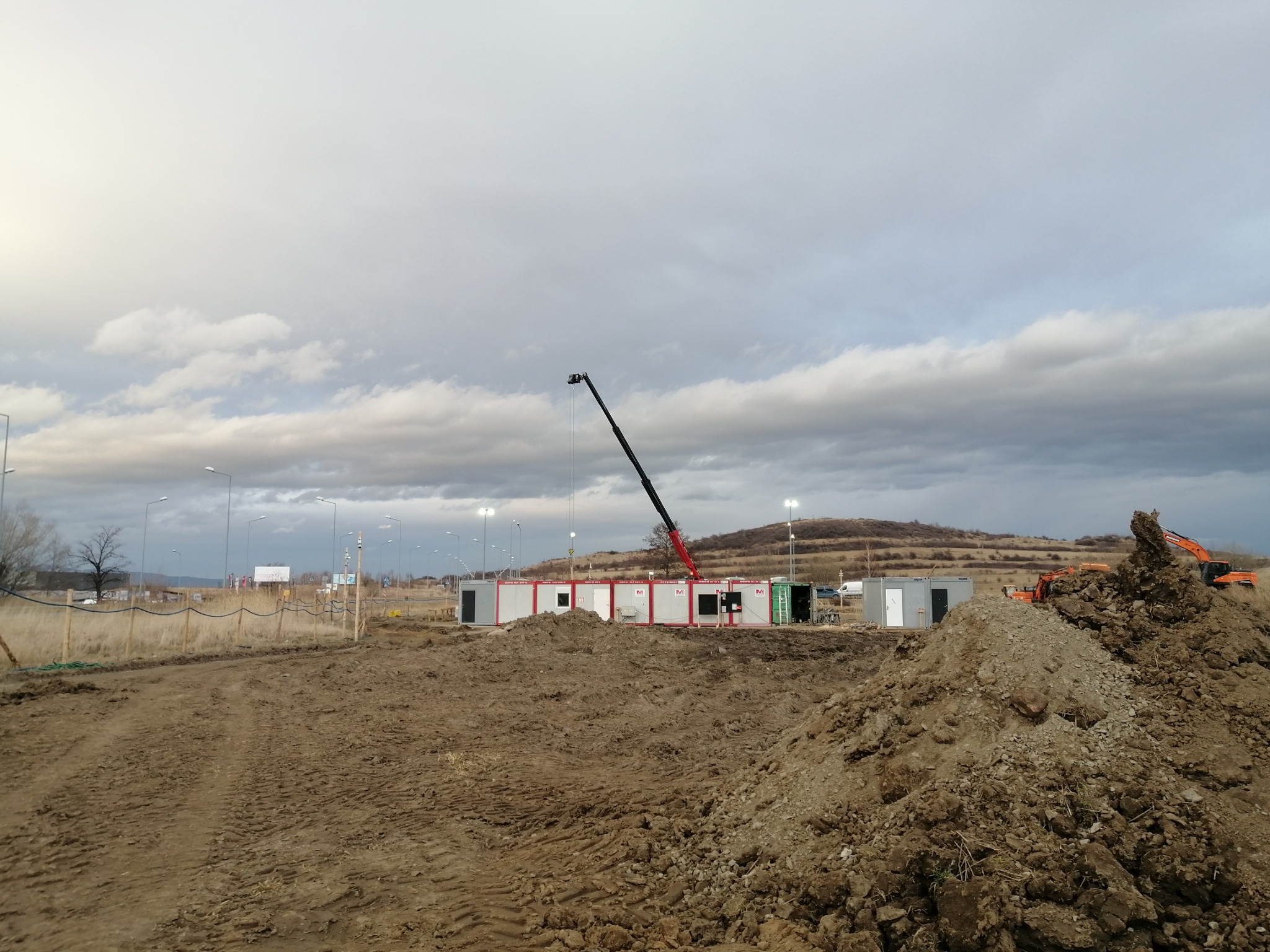 Teren budowy z kontenerami biurowymi, dźwigiem i koparką na tle pochmurnego nieba. W pierwszym planie hałda ziemi z wyraźnymi śladami opon.
