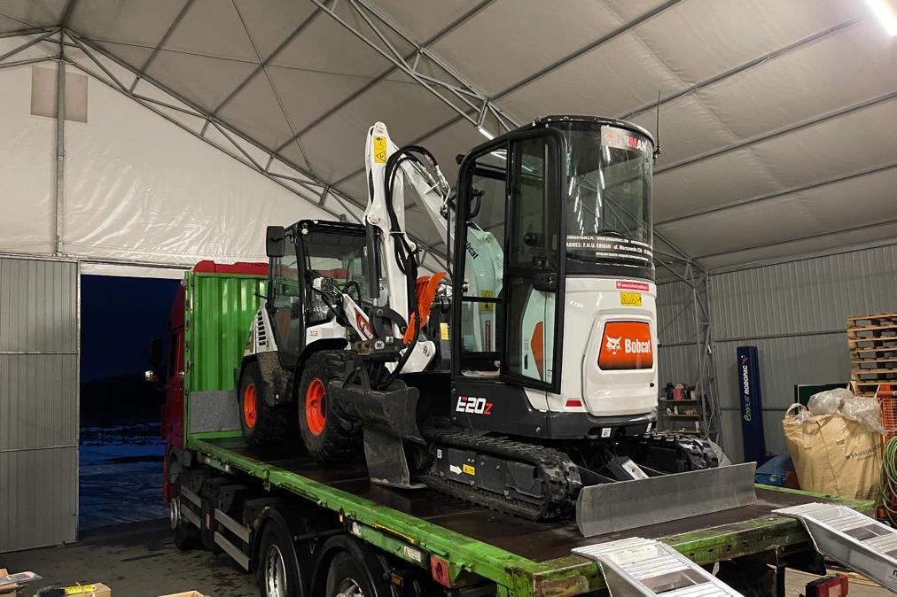 Dwie mini koparki Bobcat E20Z na zielonej platformie transportowej wewnątrz hali namiotowej. Widoczny fragment ciężarówki i palety z workami w tle.