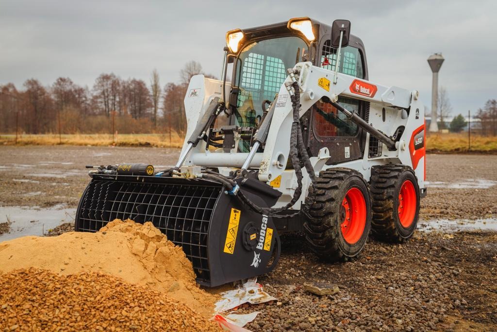 Kompaktowa ładowarka Bobcat 550 z frezem do gruntu, obok pryzma kruszywa, w tle zbiornik na wodę i drzewa bez liści.