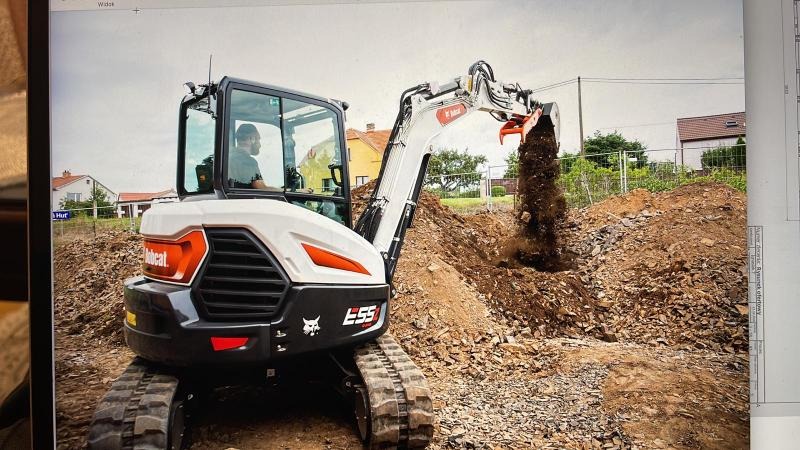 Biała mini koparka Bobcat E55 z operatorem w kabinie, podczas prac ziemnych na placu budowy; w tle widoczne domy jednorodzinne i ogrodzenie.