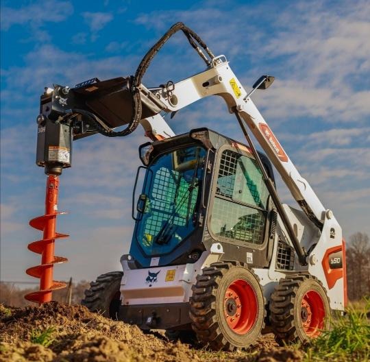 Kompaktowa ładowarka Bobcat 550 z zamontowanym wiertłem hydraulicznym, gotowa do wiercenia otworów w ziemi na tle błękitnego nieba.