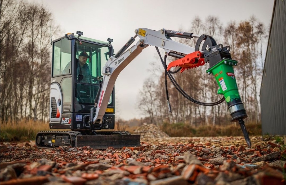 Mała koparka Bobcat E20Z z zielonym młotem hydraulicznym Montabert SC12, operator w kabinie, gruz na ziemi, budynek w tle.