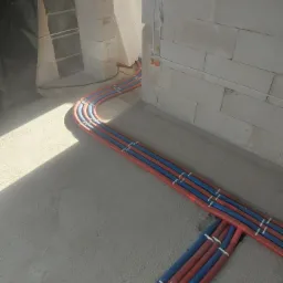 Nowa instalacja ogrzewania podłogowego z rurami w kolorze czerwonym i niebieskim, przymocowanymi do podłoża białymi zaciskami, widoczne ściany z pustaków.