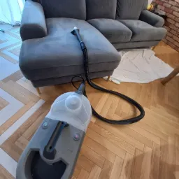 Szary narożnik z widoczną końc&oacute;wką odkurzacza piorącego na siedzisku, obok leży biała szmatka, a na pierwszym planie odkurzacz piorący na parkiecie ułożonym w jodełkę.