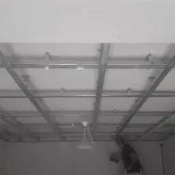 Montaż metalowej konstrukcji sufitu podwieszanego z widocznymi profilami i zawiesiami, pracownik na drabinie przykręca element do ściany.