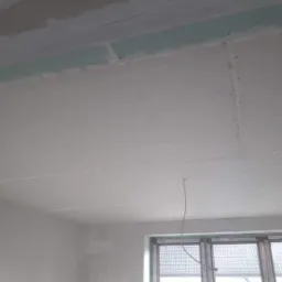 Szpachlowana powierzchnia sufitu z płyt kartonowo-gipsowych, widoczne łączenia płyt, fragment ściany i okno w tle.