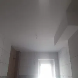 Biały sufit po remoncie, z widocznym miejscem na lampę i fragmentem dekoracyjnego obniżenia sufitu, oraz fragmentem ściany z białymi płytkami i oknem.