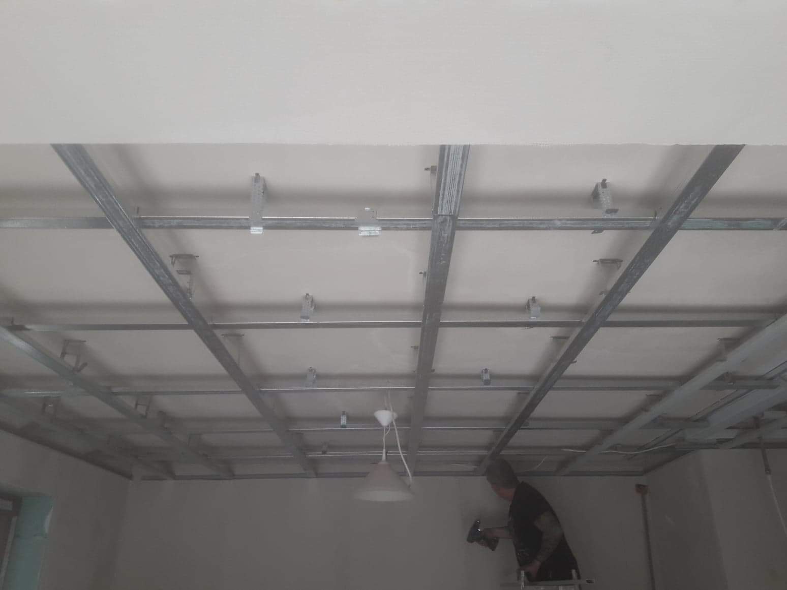 Montaż metalowej konstrukcji sufitu podwieszanego z widocznymi profilami i zawiesiami, pracownik na drabinie przykręca element do ściany.