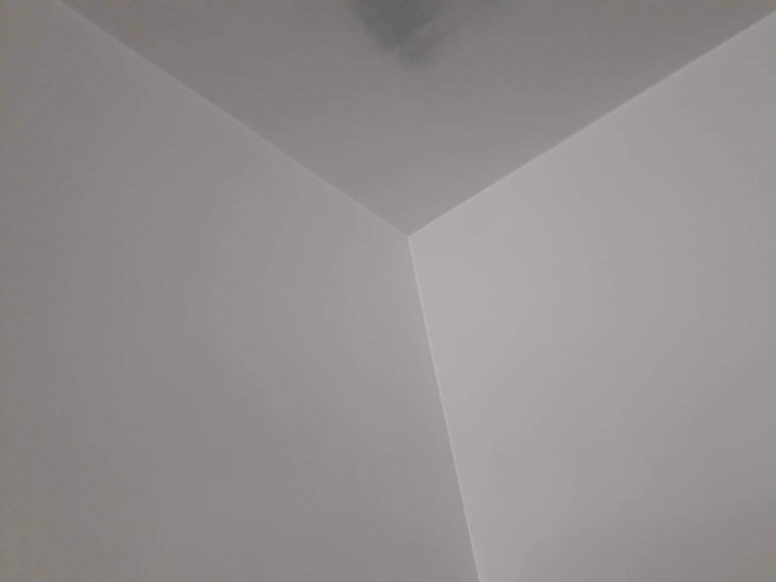 Pomieszczenie po świeżym malowaniu, widoczny fragment rogu ściany i sufitu, jednolity jasny odcień farby na całej powierzchni, minimalne niedoskonałości w górnym rogu sufitu.