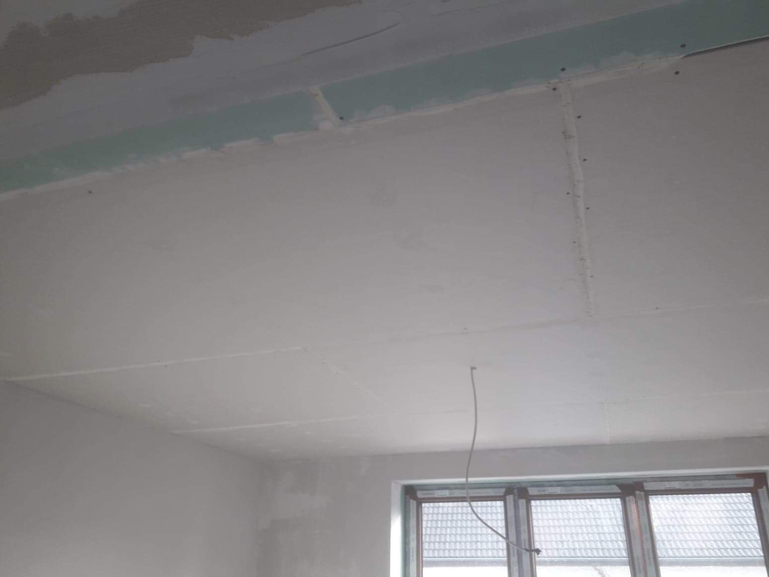 Szpachlowana powierzchnia sufitu z płyt kartonowo-gipsowych, widoczne łączenia płyt, fragment ściany i okno w tle.