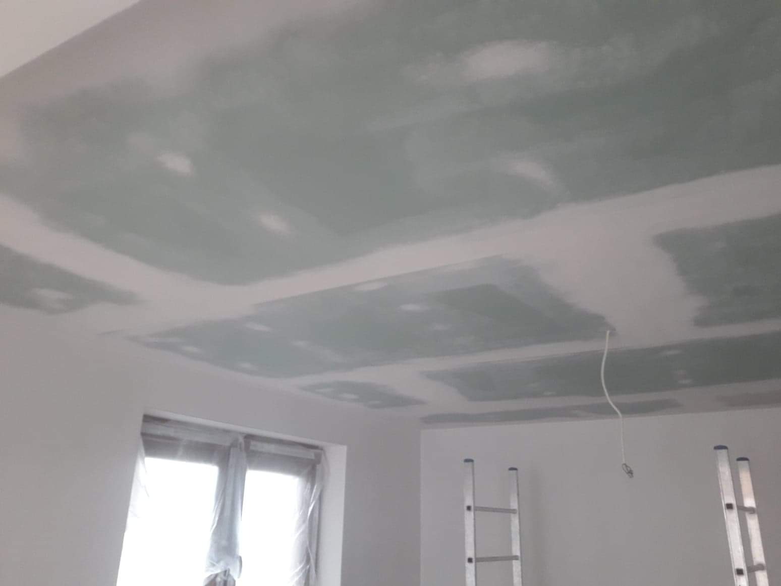 Sufit z płyt kartonowo-gipsowych w trakcie szpachlowania, widoczne łączenia płyt i wiszący kabel, drabina oparta o ścianę, okno zabezpieczone folią.