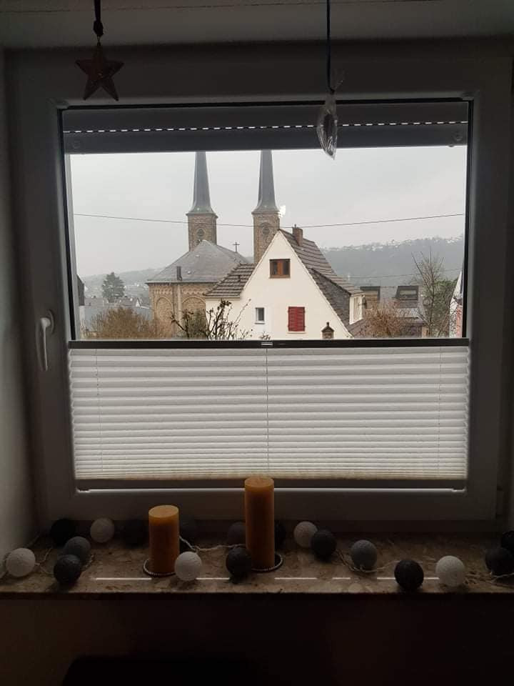 Biała plisa okienna w połowie zasłonięta, na parapecie dekoracja ze świec i cotton balls, w tle widok na kościół i zabudowania.