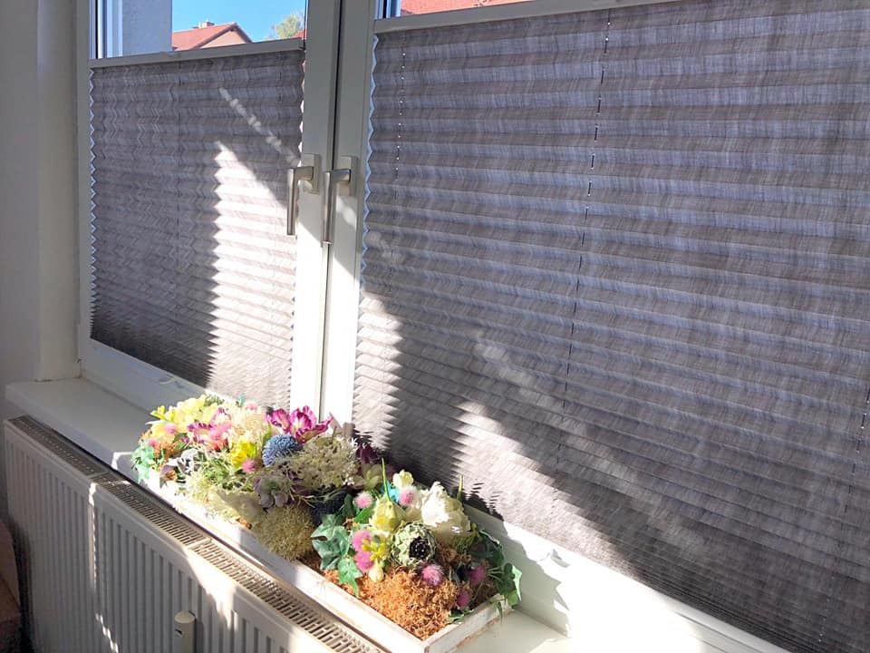 Szare, plisowane osłony okienne w oknie z dekoracją kwiatową na parapecie, oświetlone popołudniowym słońcem.