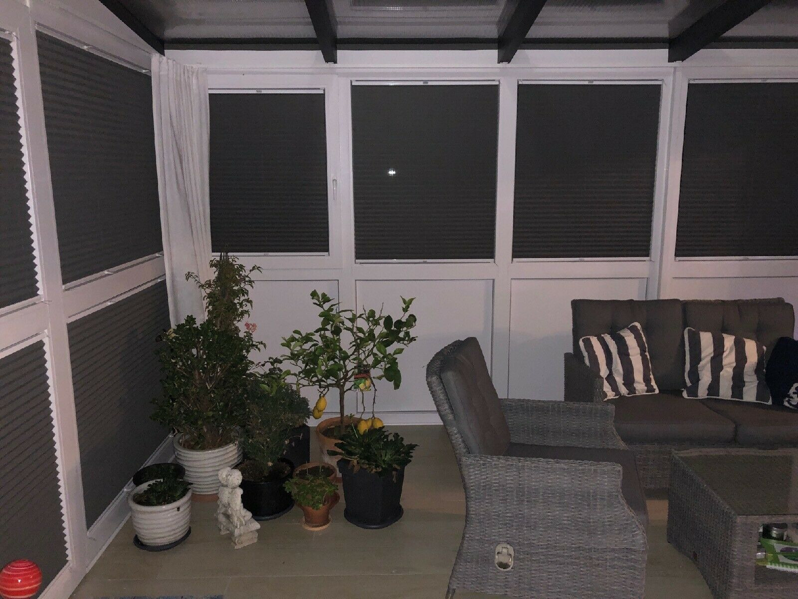 Zaciemnione wnętrze z plecionymi meblami ogrodowymi i roślinami doniczkowymi przed oknami z plisowanymi osłonami.