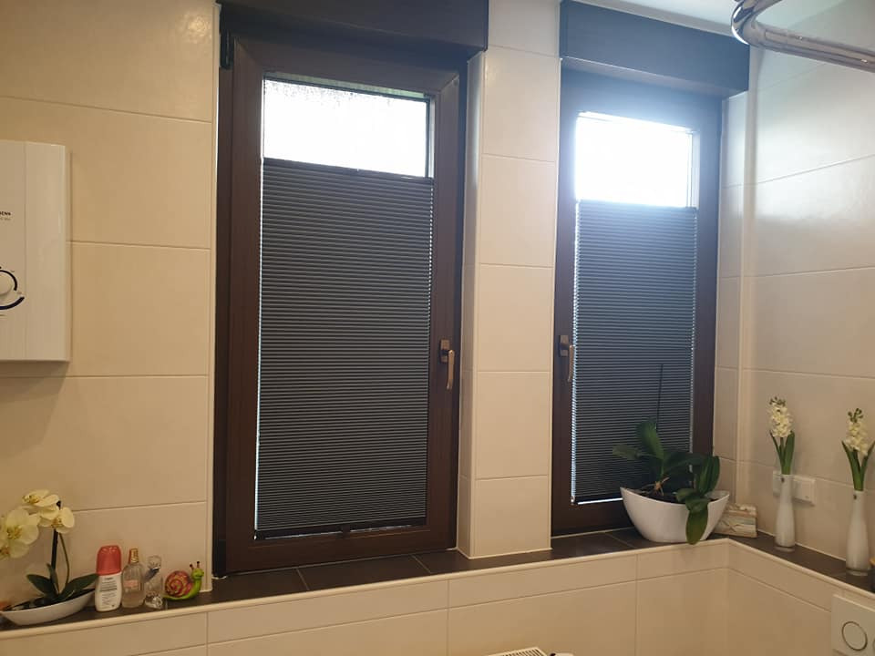 Dwa okna w łazience w Łodzi z zamontowanymi szarymi plisami, widoczne dekoracje okienne i rośliny.