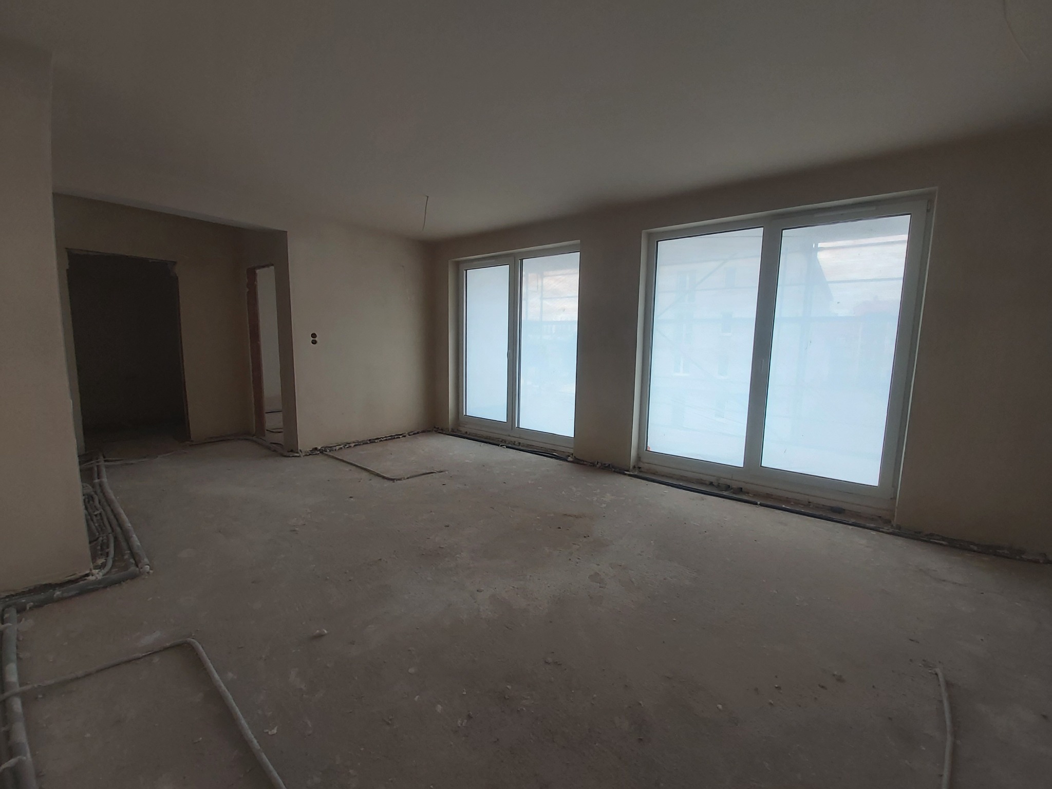 Wnętrze w Rybniku przed pracami wykończeniowymi: surowy stan, widoczne instalacje elektryczne przy podłodze, duże okna z białymi ramami, ściany przygotowane do dalszych prac.