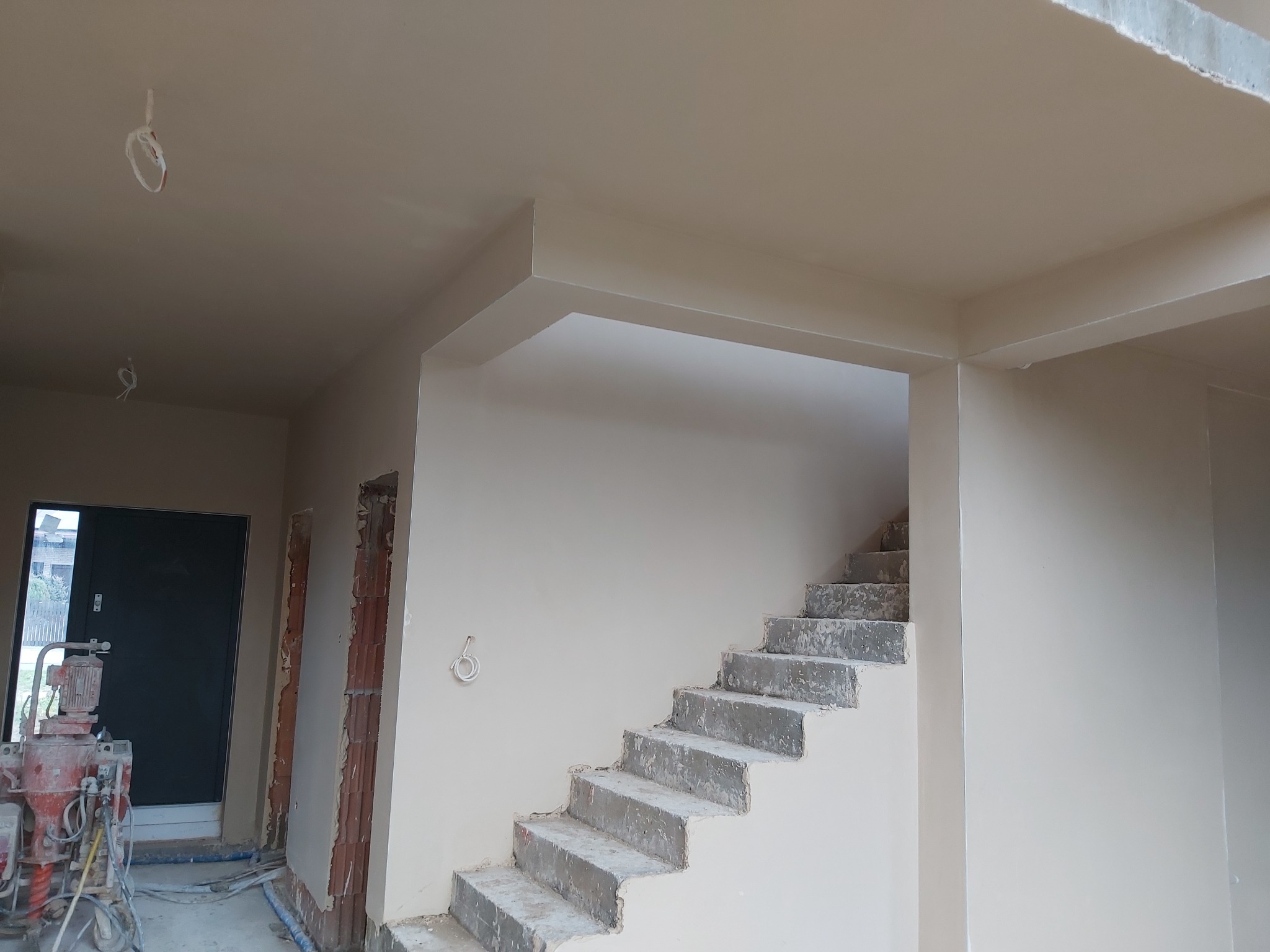 Wnętrze domu w trakcie prac wykończeniowych: świeżo położone gładzie na ścianach i suficie, surowe betonowe schody prowadzące na piętro, widoczna maszyna do nakładania tynków przy wejściu.