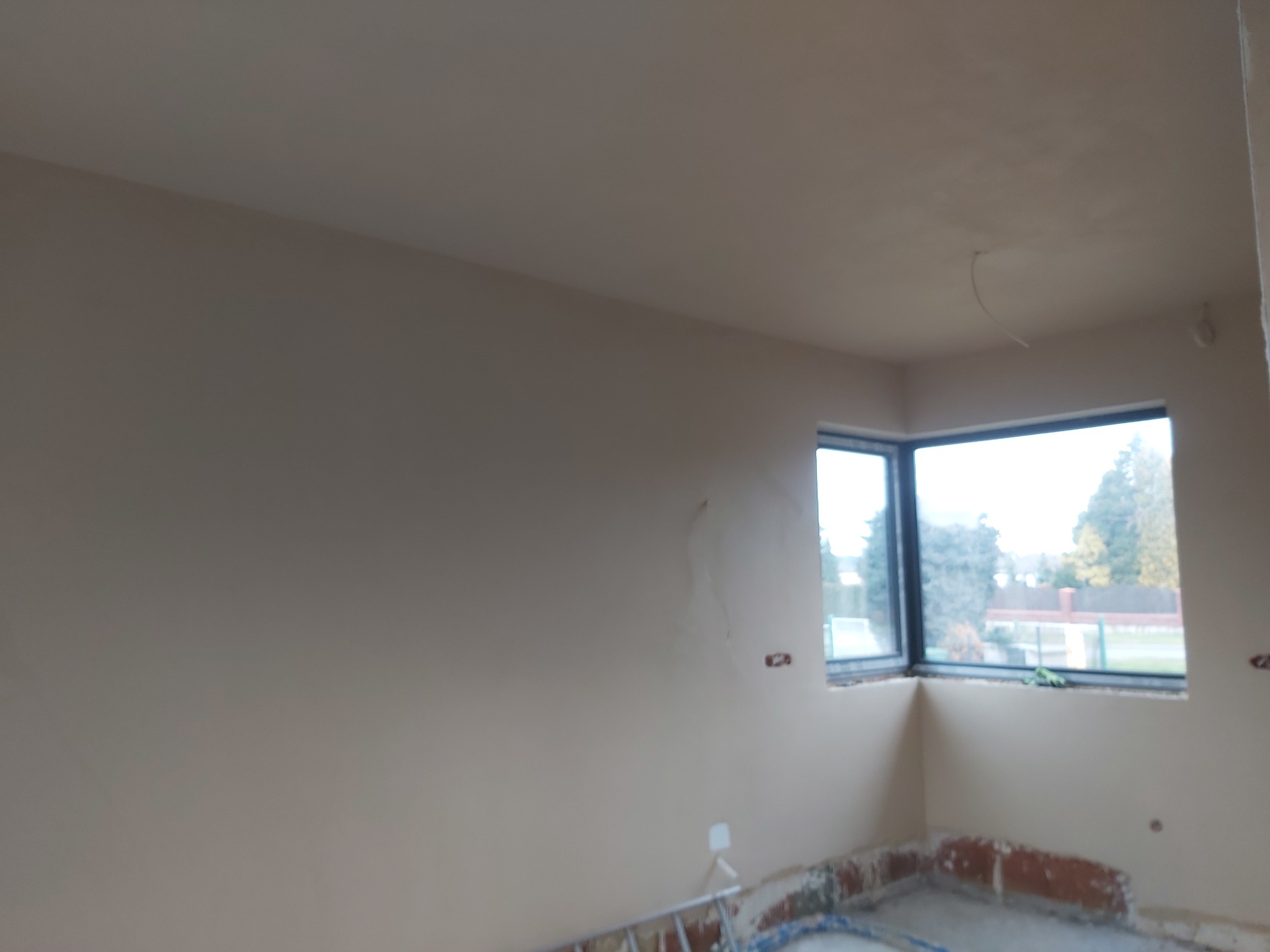 Wnętrze pokoju po nałożeniu jasnej, gładkiej warstwy tynku, widoczne niedoskonałości i ślady po instalacjach, okno narożne z widokiem na zewnątrz.