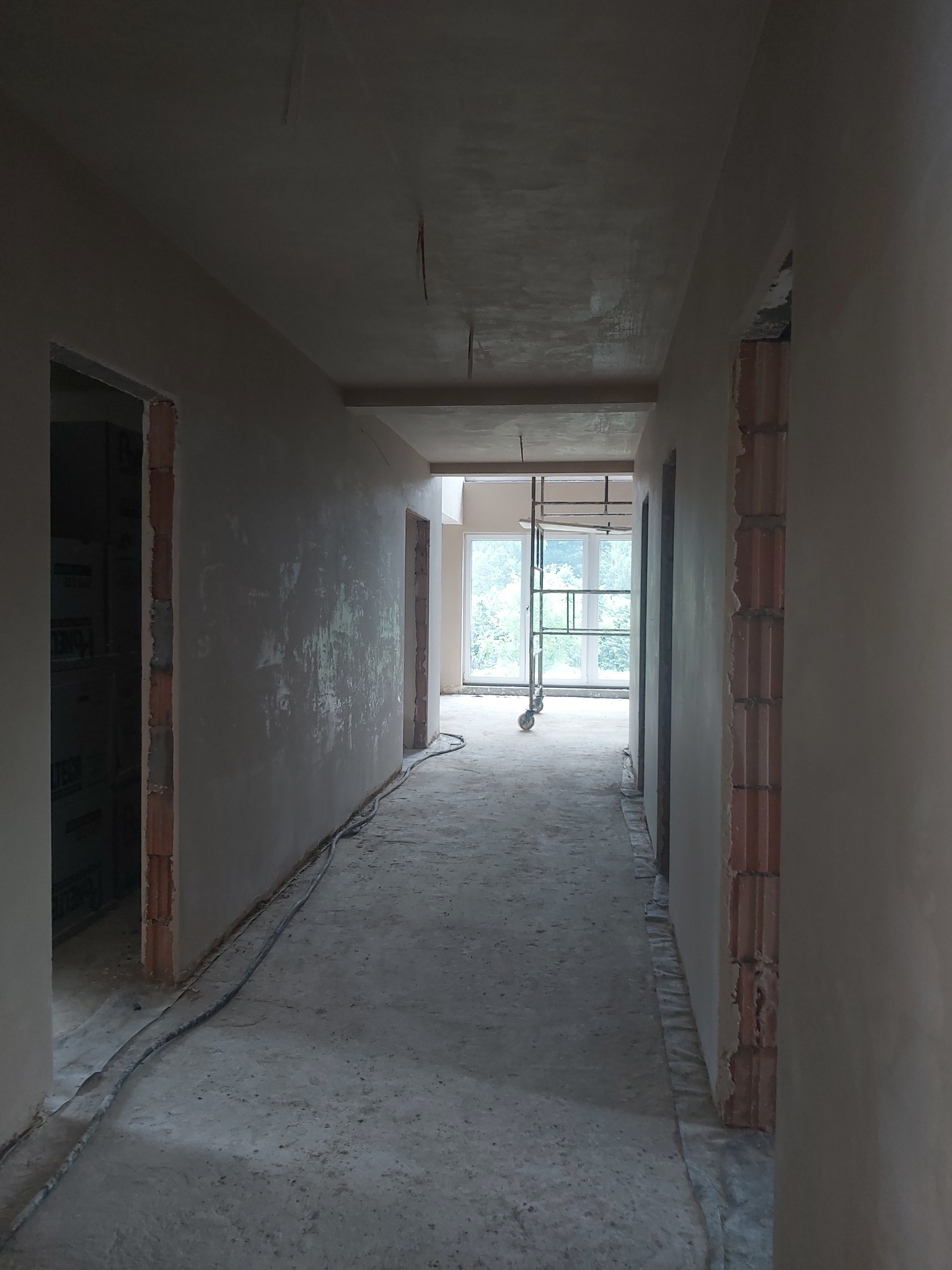 Korytarz w trakcie prac wykończeniowych, ściany pokryte surowym tynkiem, widoczne fragmenty cegieł wokół ościeżnic, na podłodze leżą przewody, w oddali rusztowanie.
