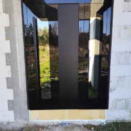 Montaż czarnych drzwi zewnętrznych z klejonym szkłem, widoczne taśmy izolacyjne, surowy stan budynku w tle.