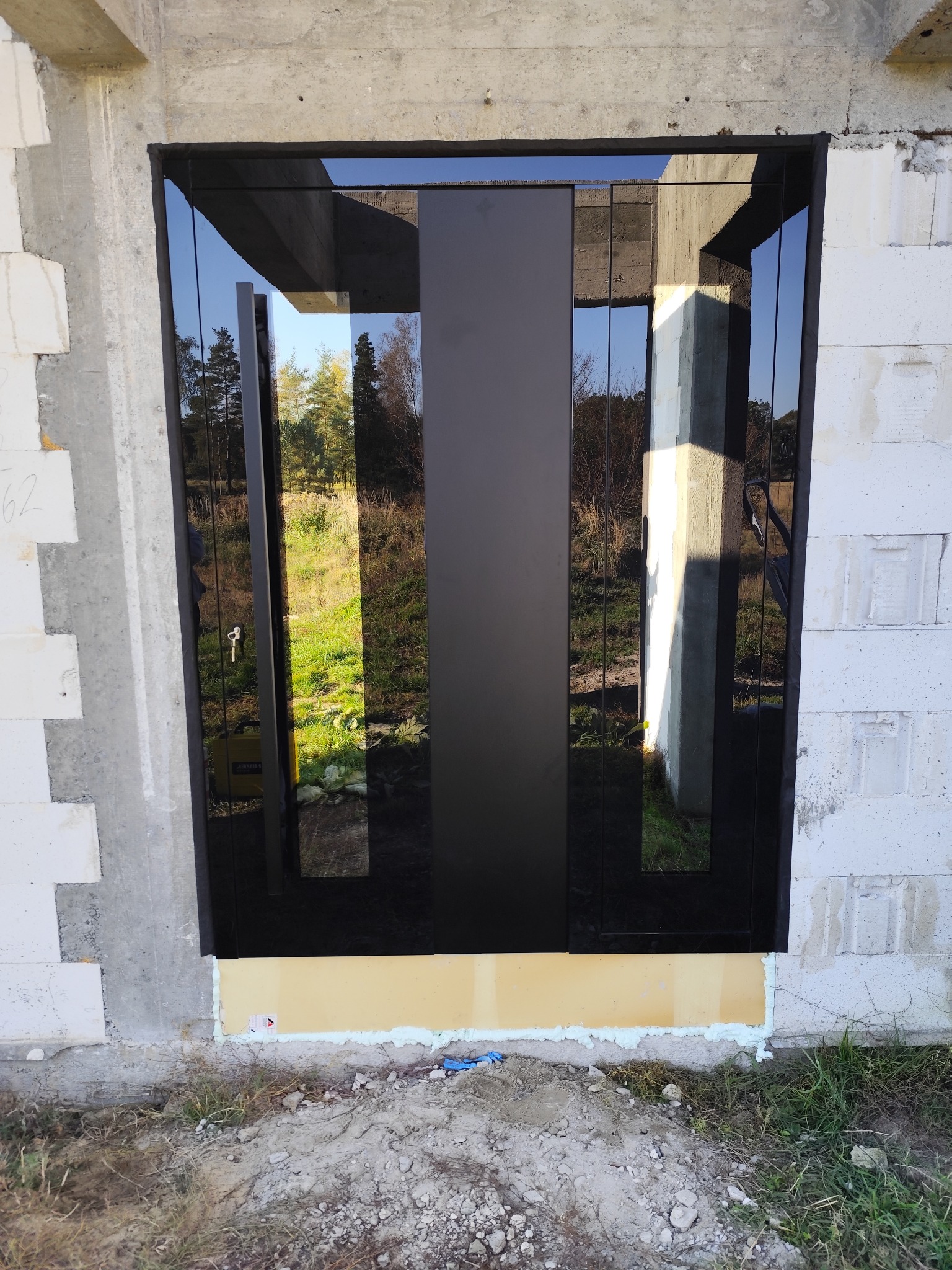 Montaż czarnych drzwi zewnętrznych z klejonym szkłem, widoczne taśmy izolacyjne, surowy stan budynku w tle.