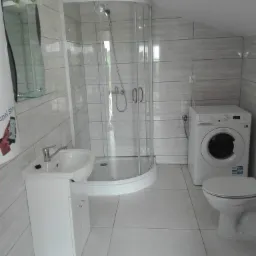 Ukończona łazienka w odcieniach szarości i bieli z kabiną prysznicową narożną, umywalką z szafką, pralką i toaletą.