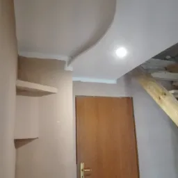 Wnętrze w trakcie prac wykończeniowych: sufit z dekoracyjnym zaokrągleniem, fragment drewnianych schodów, drzwi z klamką i półki w ścianie przed malowaniem.