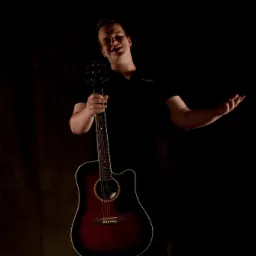 Młody mężczyzna w czarnej koszulce uśmiecha się, trzymając akustyczną gitarę z czerwono-czarnym gradientem na tle ciemnego, teksturowanego tła.