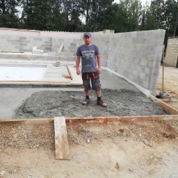 Pracownik w szarym t-shircie i krótkich spodniach na tle wylewki betonowej, częściowo ułożonej w szalunku, z widocznymi ścianami z bloczków betonowych w tle.