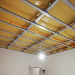 Stelaż sufitu podwieszanego z izolacją z folii, widoczne profile metalowe i pomarańczowe przewody, w tle drabiny i drzwi