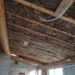 Poddasze z drewnianą konstrukcją, ocieplone wełną mineralną, widoczne przewody elektryczne i fragment ściany z pustaków.