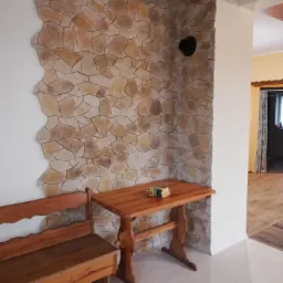 Fragment wnętrza z dekoracyjną ścianą wyłożoną kamieniem, drewnianym stołem z paczką sera i ławką, widok na sąsiednie pomieszczenie z zasłoną w oknie.