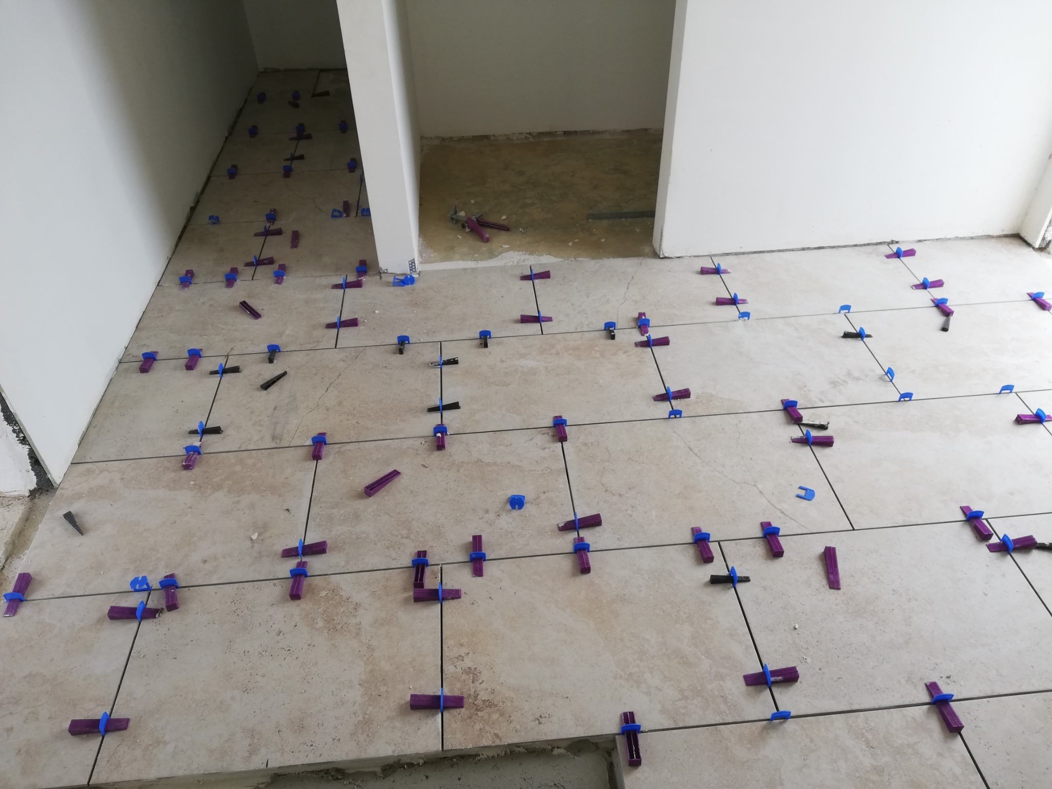 Układanie beżowych płytek podłogowych z użyciem poziomujących klipsów i klinów w pomieszczeniu z białymi ścianami i otwartym przejściem.