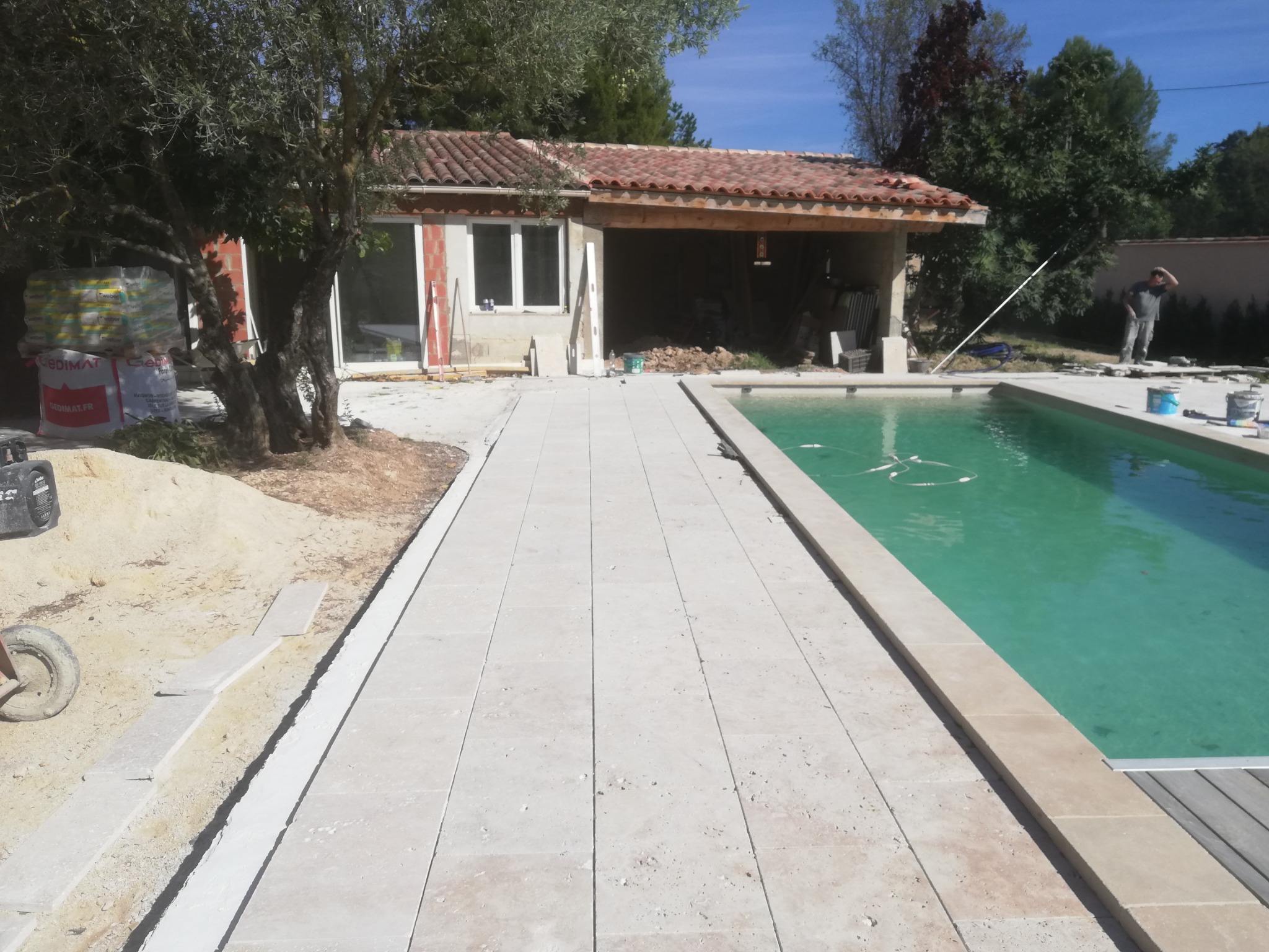 Układanie jasnych, prostokątnych płyt chodnikowych wokół basenu, widoczna konstrukcja budynku z czerwonym dachem w tle, prace w toku.