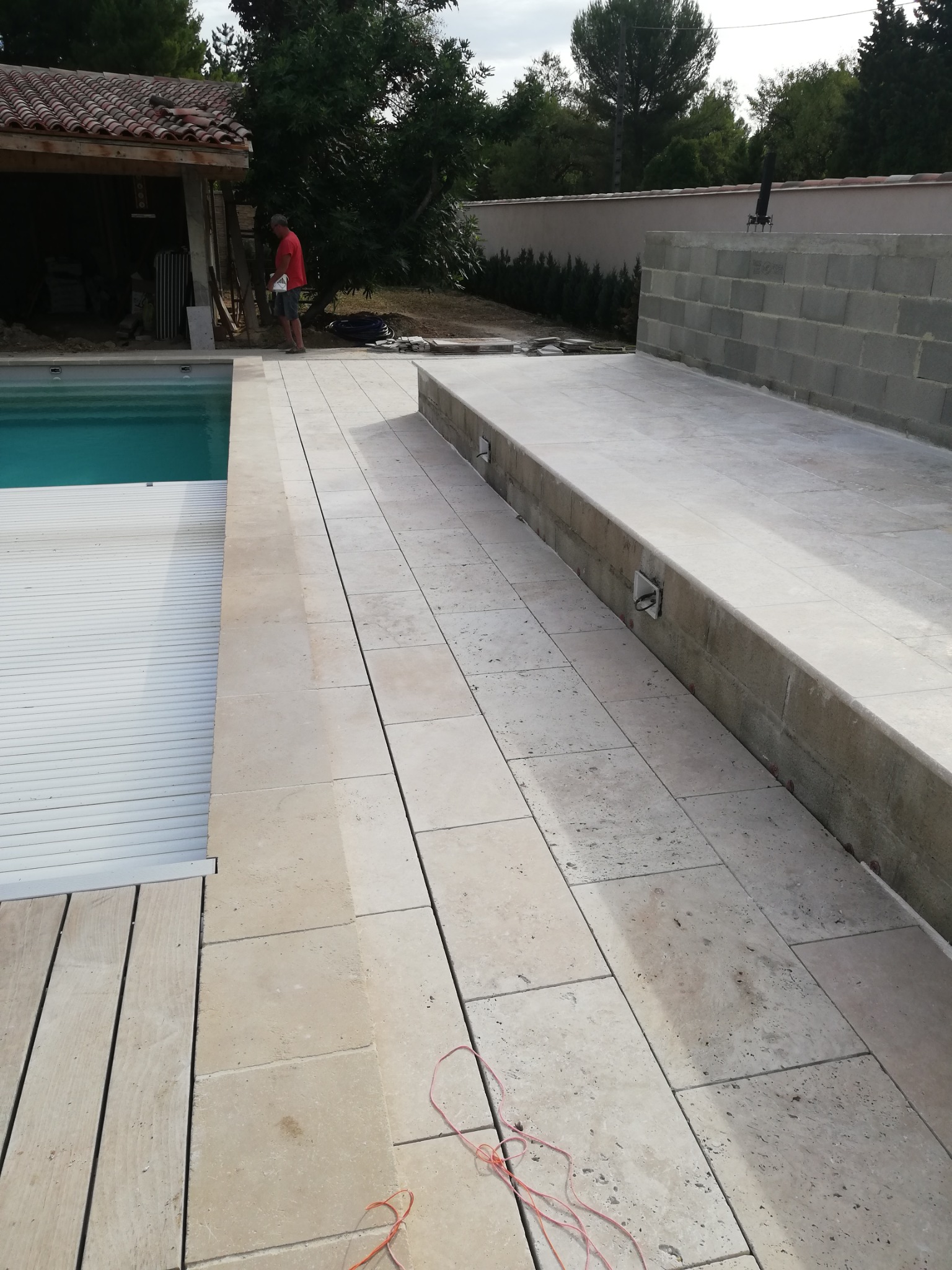 Jasne płytki trawertynowe układane wokół basenu z automatyczną roletą, widoczny fragment ściany z bloczków i pracownik w oddali.