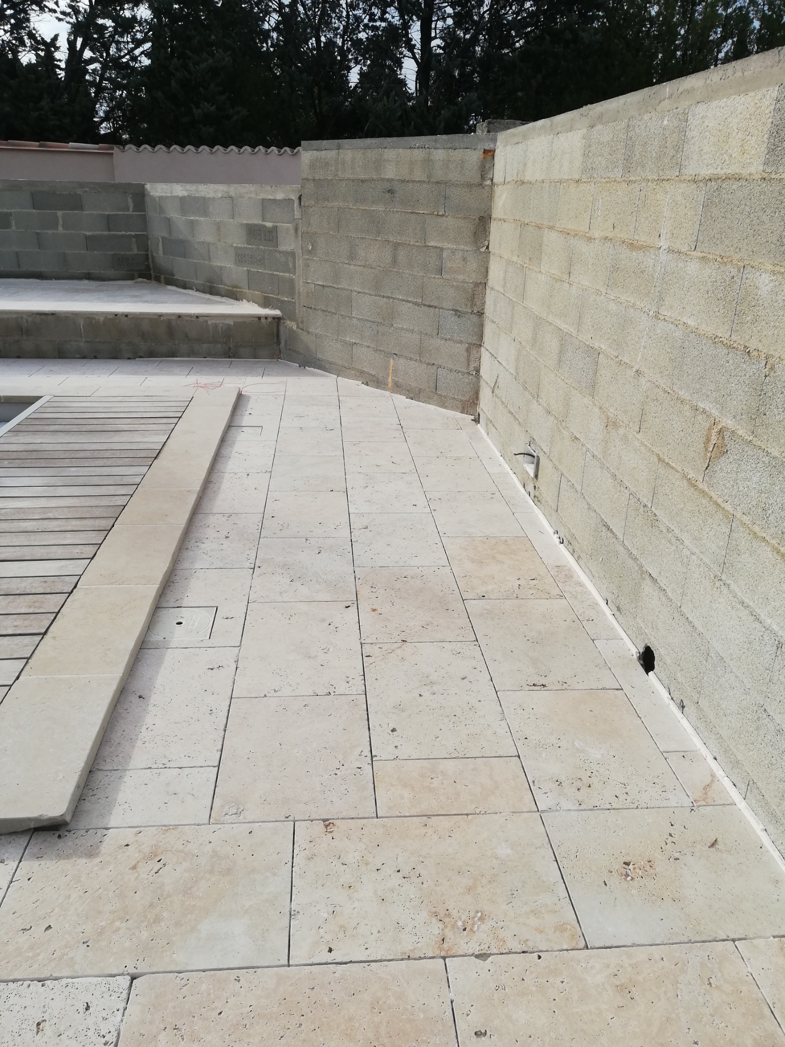 Układanie jasnych, trawertynowych płytek na tarasie z surowymi ścianami z bloczków betonowych w tle.