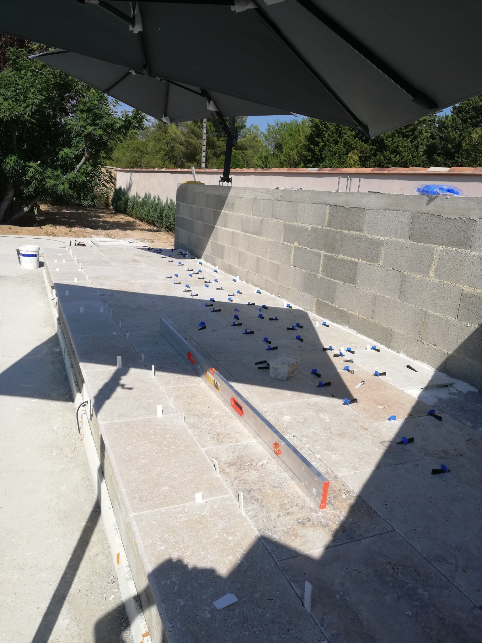 Układanie jasnych, kwadratowych płytek na zewnątrz budynku, z użyciem poziomicy i niebieskich klinów dystansowych, widoczne bloczki betonowe w tle, zacienione przez duży parasol.