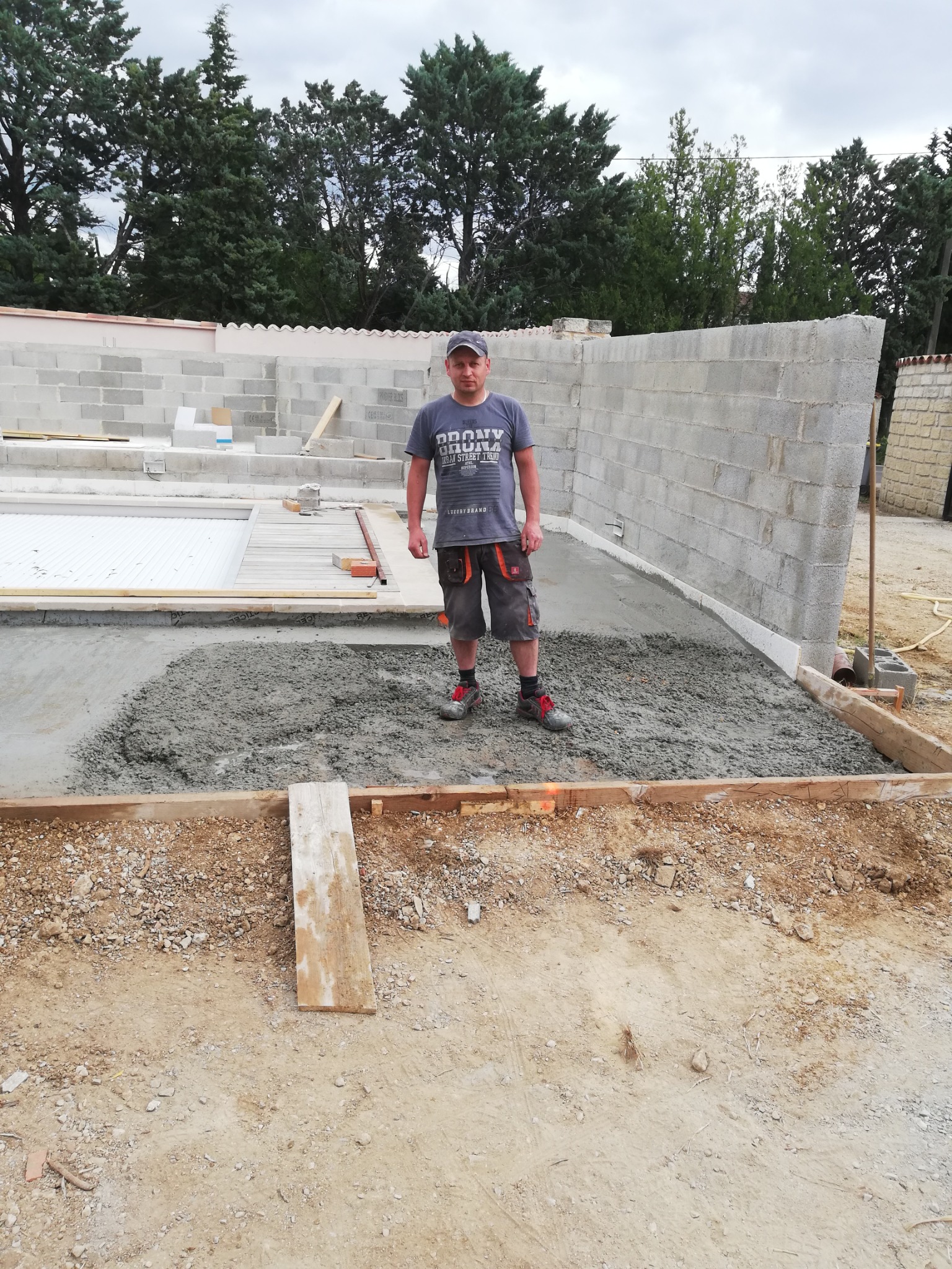 Pracownik w szarym t-shircie i krótkich spodniach na tle wylewki betonowej, częściowo ułożonej w szalunku, z widocznymi ścianami z bloczków betonowych w tle.