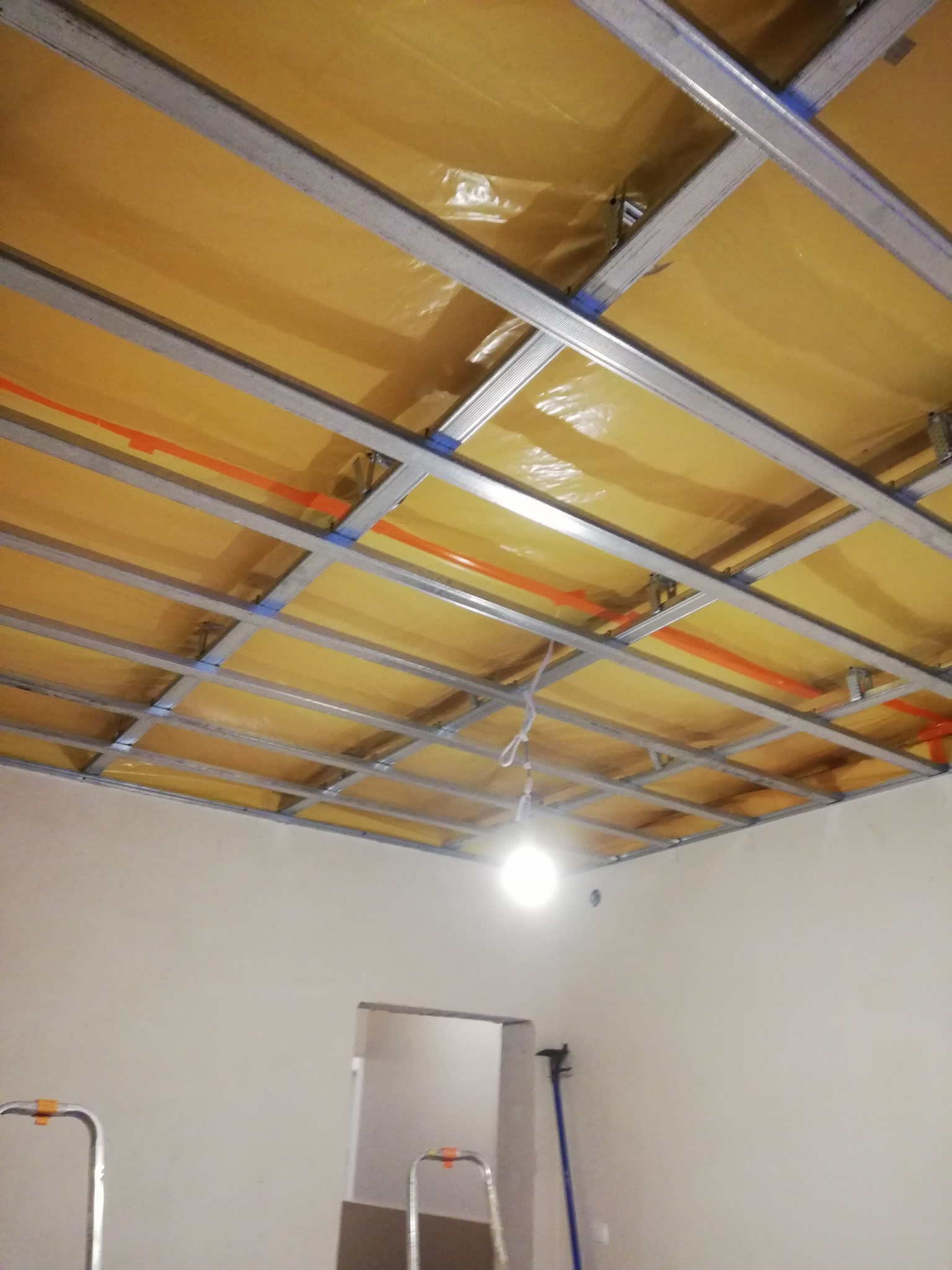 Stelaż sufitu podwieszanego z izolacją z folii, widoczne profile metalowe i pomarańczowe przewody, w tle drabiny i drzwi