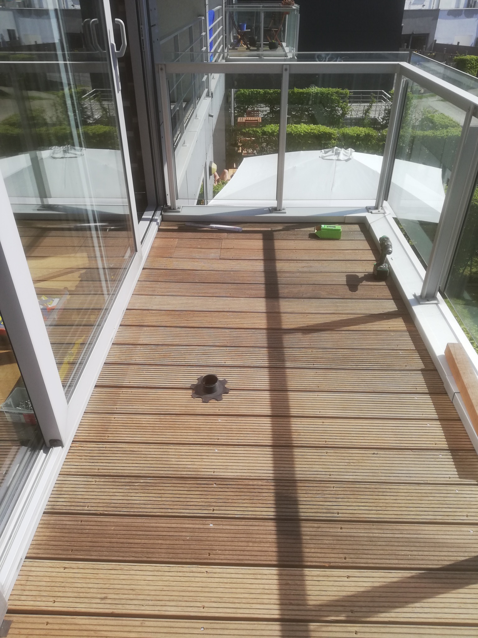 Drewniany taras na balkonie z widocznymi śrubami, narzędziem i elementem mocującym, częściowo osłonięty szklaną balustradą i oświetlony naturalnym światłem.