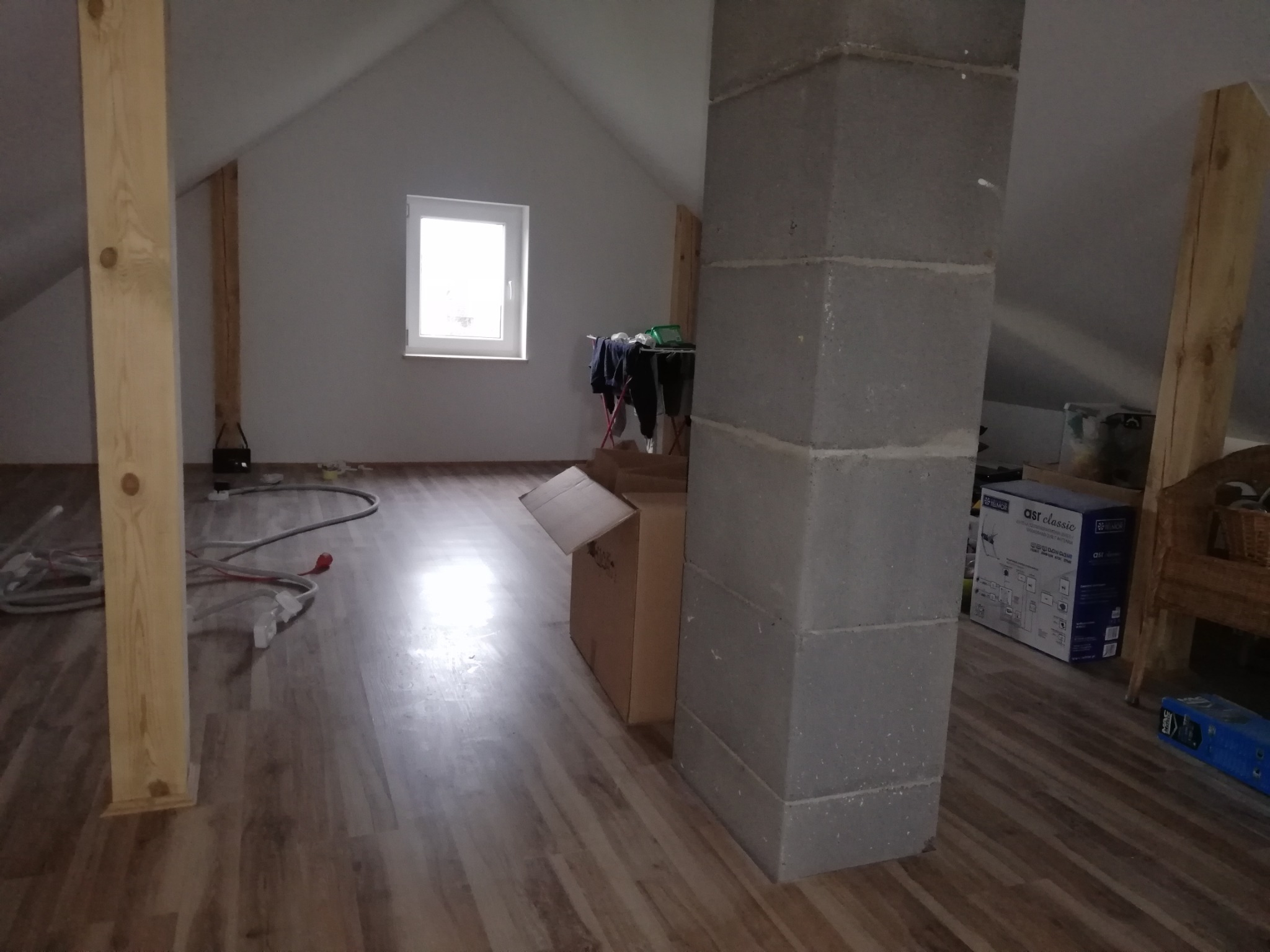 Poddasze w trakcie remontu z widoczną konstrukcją drewnianą, oknem, pionem z bloczków betonowych, częściowo ułożoną podłogą z paneli, kablami i kartonami.
