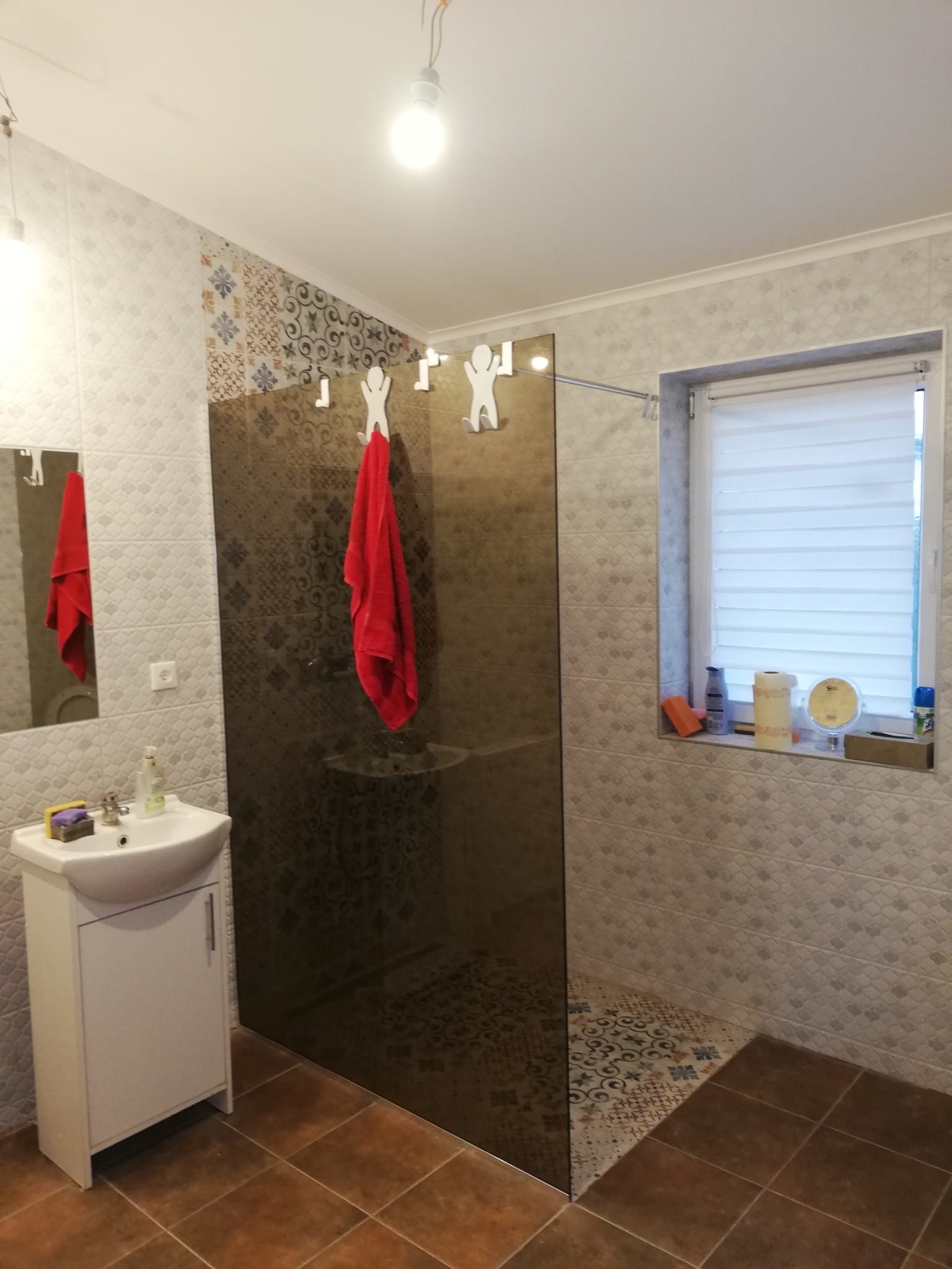 Umywalka z szafką, lustro i kabina prysznicowa z brązowego szkła w wykończonej łazience z beżowymi płytkami i dekoracyjnymi wzorami, czerwony ręcznik na wieszaku.