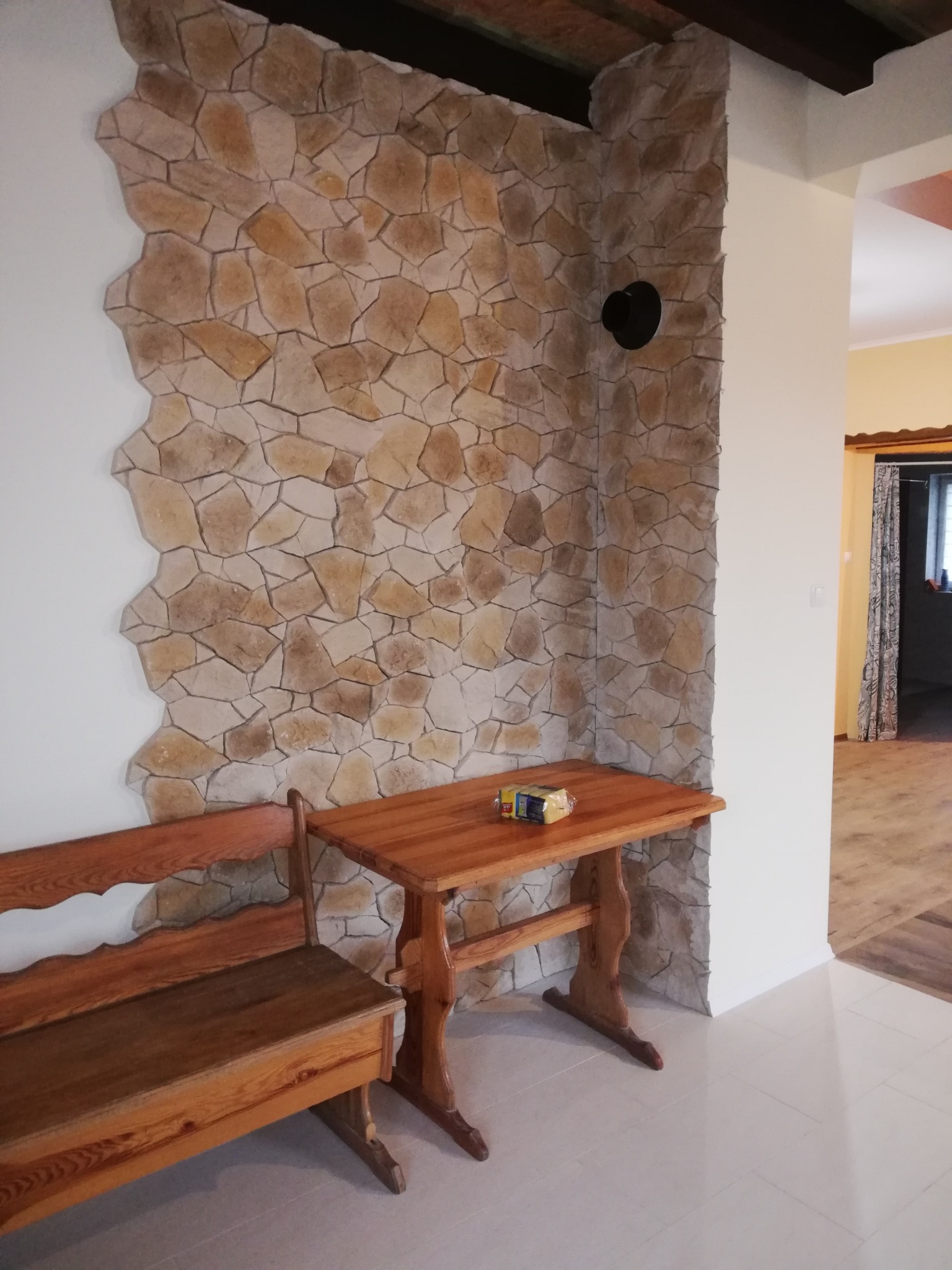 Fragment wnętrza z dekoracyjną ścianą wyłożoną kamieniem, drewnianym stołem z paczką sera i ławką, widok na sąsiednie pomieszczenie z zasłoną w oknie.