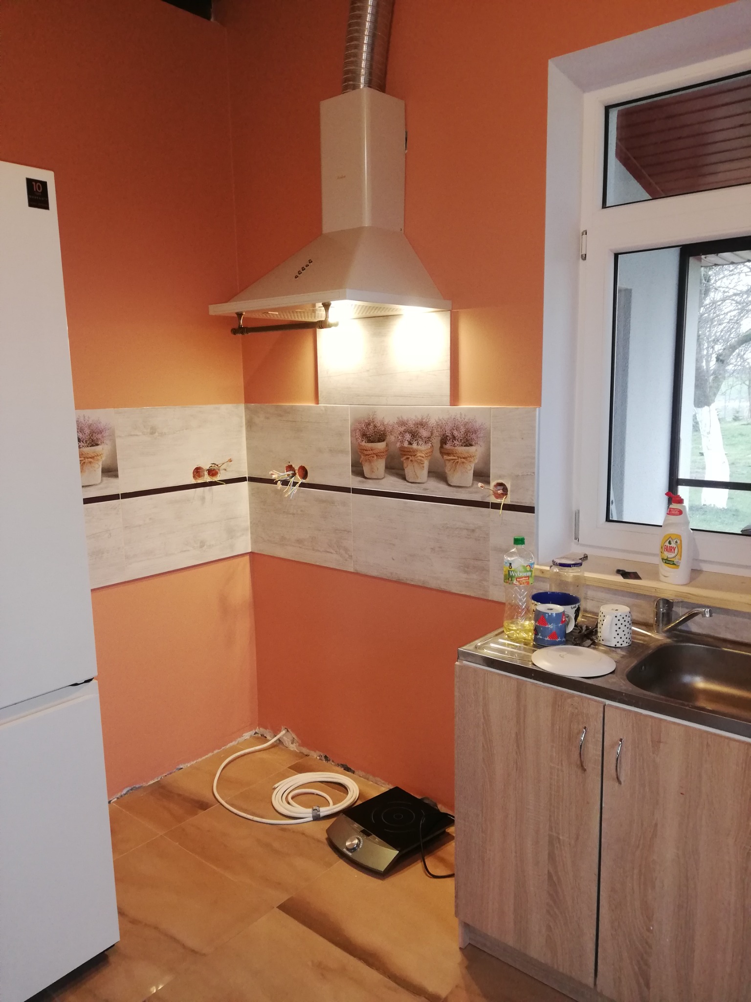 Kuchnia w trakcie remontu: pomarańczowe ściany, częściowo ułożone płytki z motywem lawendy, biały okap, biała lodówka i szafka pod zlewem z drewnianym wykończeniem. Na podłodze leży zwinięty kabel...