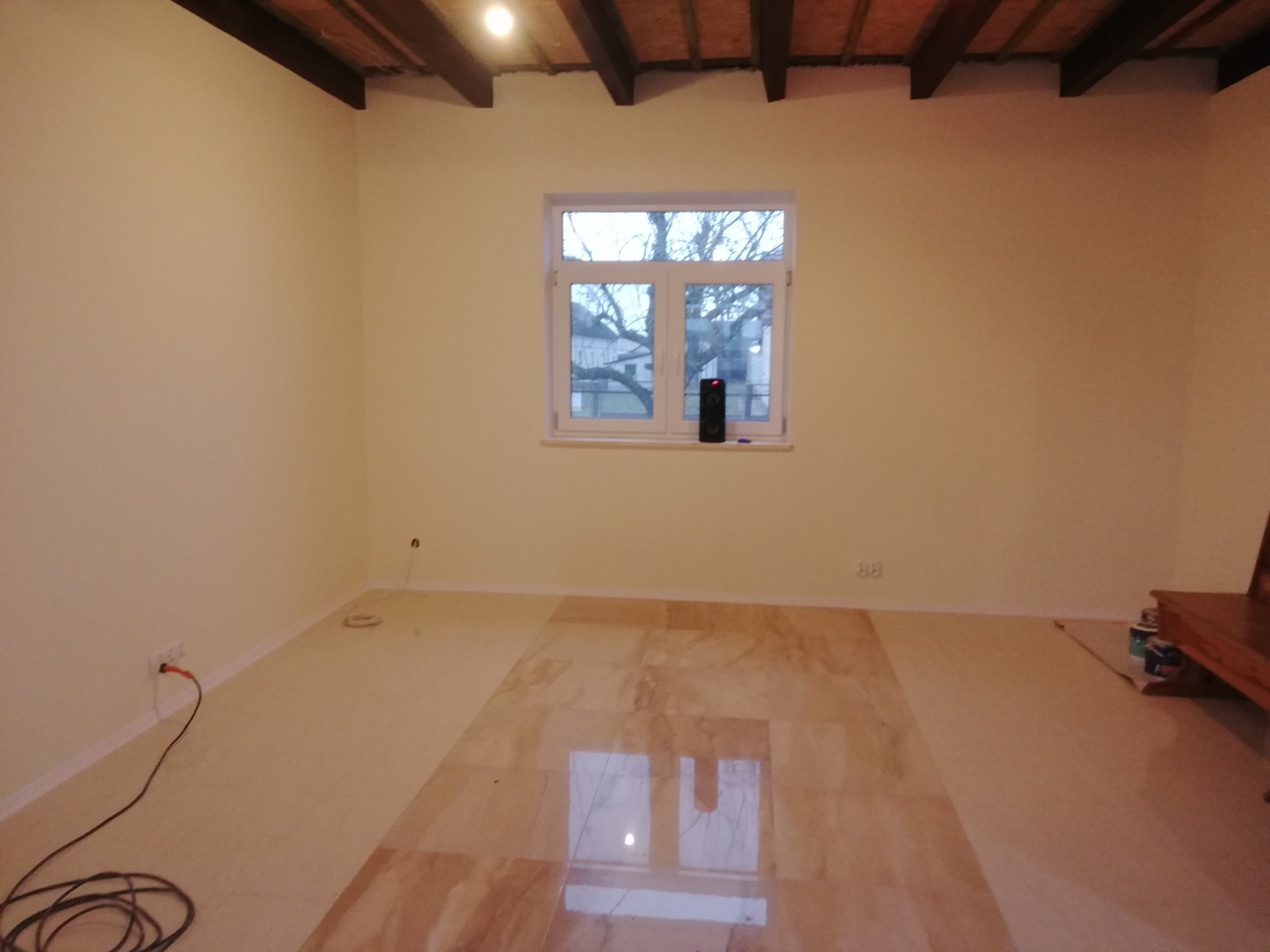 Wnętrze pokoju po remoncie: świeżo pomalowane ściany w odcieniach beżu, błyszcząca podłoga z jasnych paneli odbijająca światło z okna, widoczne drewniane belki stropowe, gniazdko elektryczne...