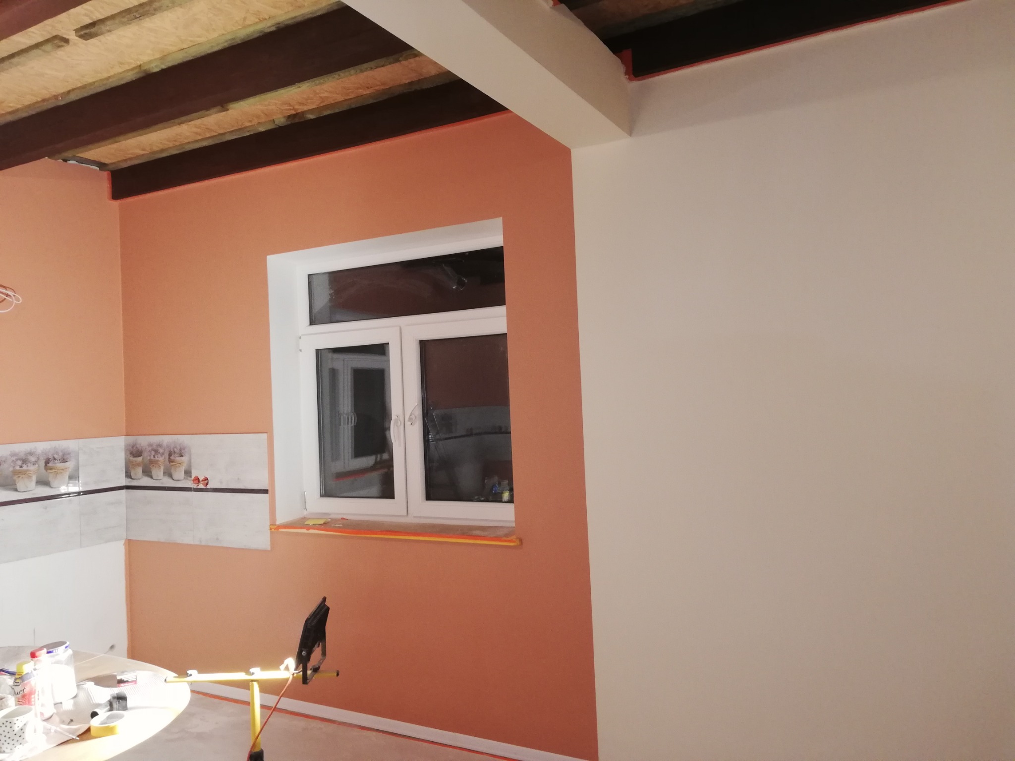Wnętrze w trakcie malowania: ściana pomalowana na pomarańczowo, biała ściana obok okna, widoczne narzędzia malarskie i zabezpieczenia, fragment sufitu z drewnianymi elementami.